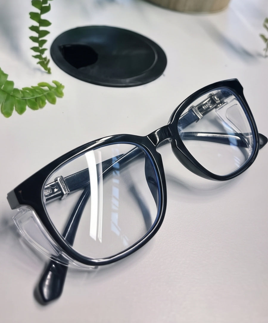 Adapt Dew glasses in black, close-up on a desk with a green plant in the background